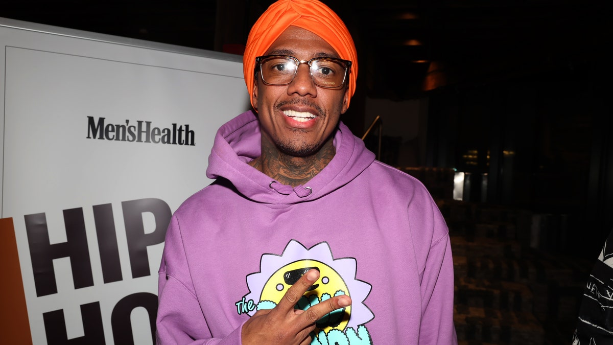 Nick Cannon in a purple sweatshirt throwing up a piece sign and wearing a red cap