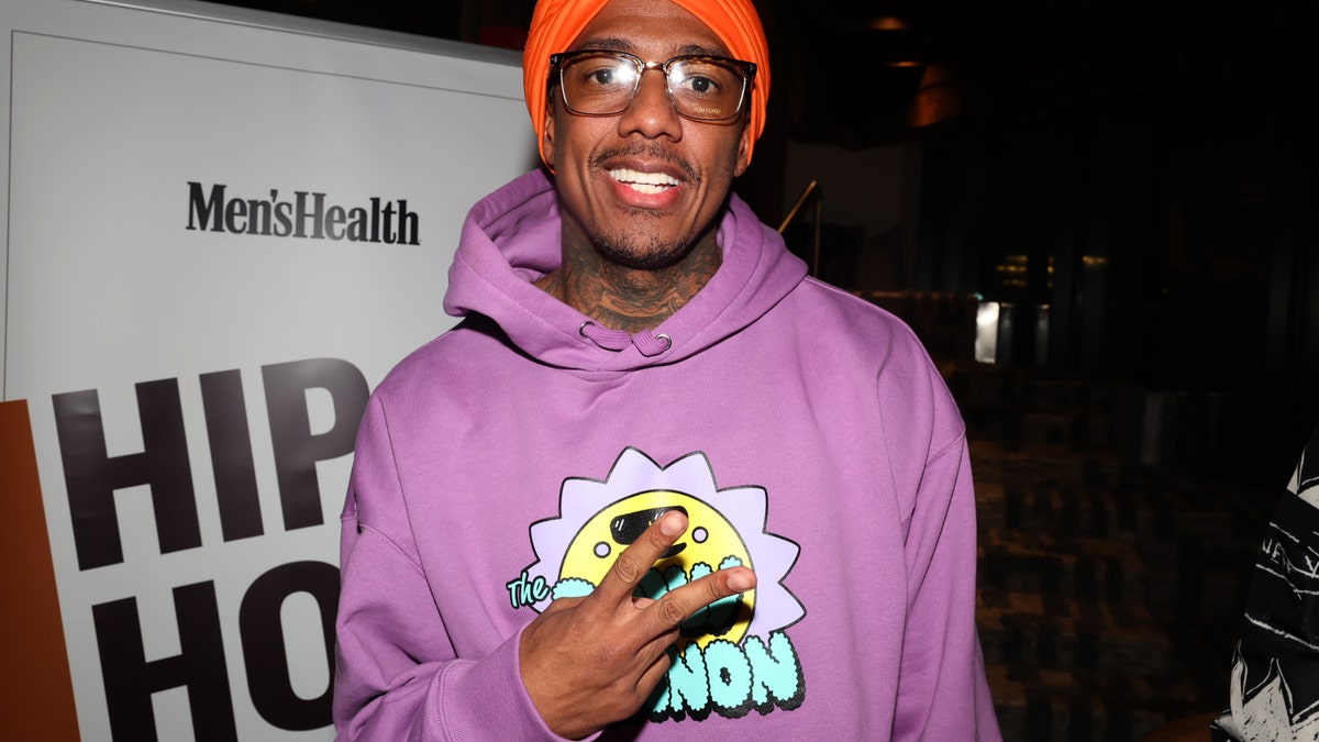 Nick Cannon in a purple sweatshirt throwing up a piece sign and wearing a red cap