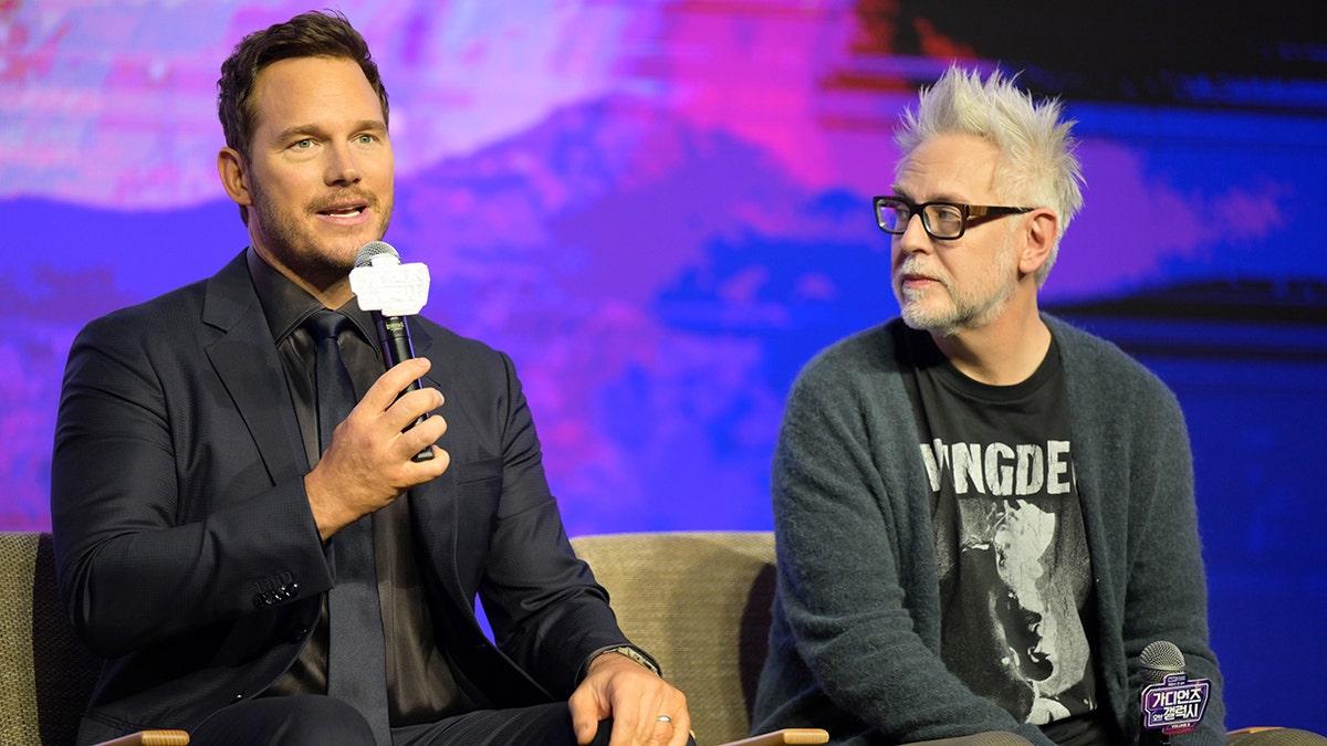 Chris Pratt wearing a blue suit and black shirt speaks into the microphone, sitting next to director James Gunn who is wearing a black t-shirt and blue sweater on stage