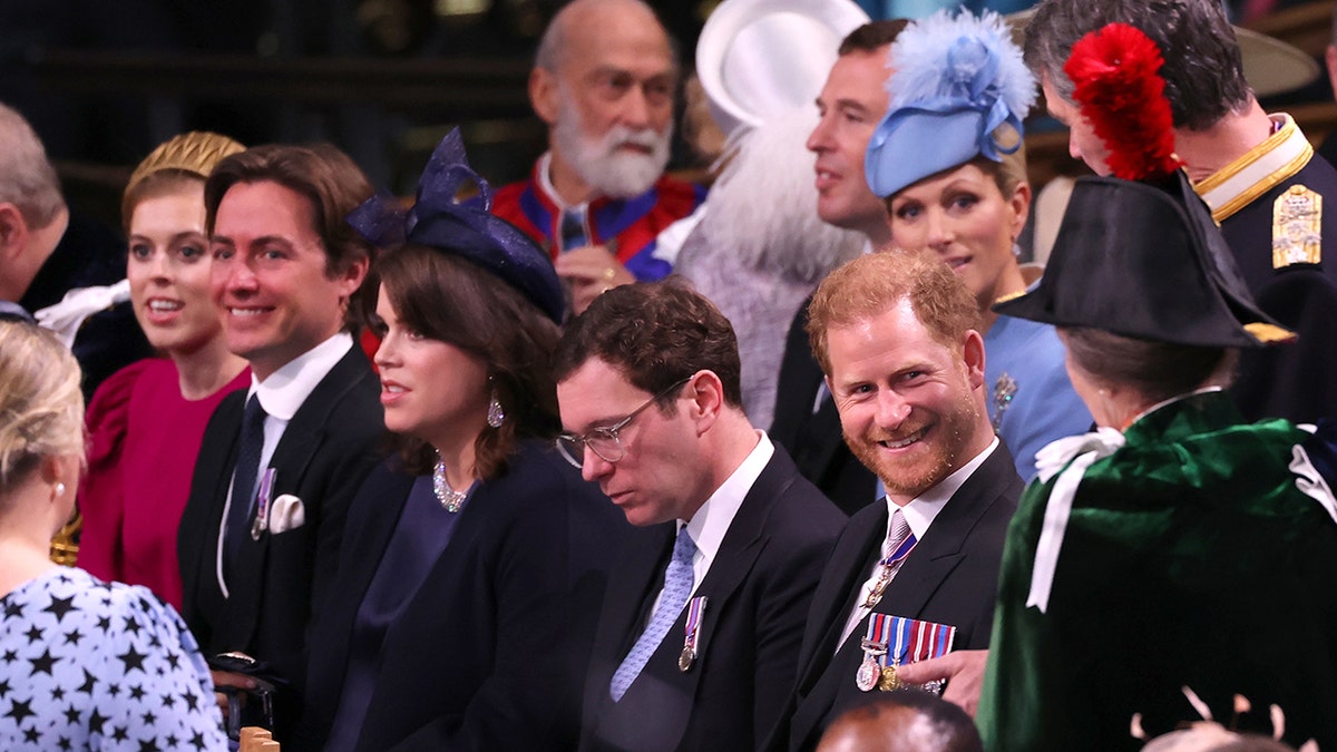 Prince Harry, seated, smiles in a suit as his aunt Anne, Princess Royal approaches him wearing a green jacket and dark hat with a red feather in it (with her back to the camera)