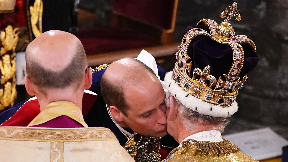 Prince William kisses his father's left cheek, who is sitting in a chair with a dark crown as he is crowned King