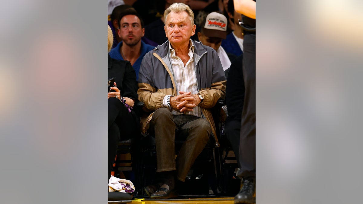 Pat Sajak sits courtside in a two-toned blue and tan raincoat with his hands clasped together