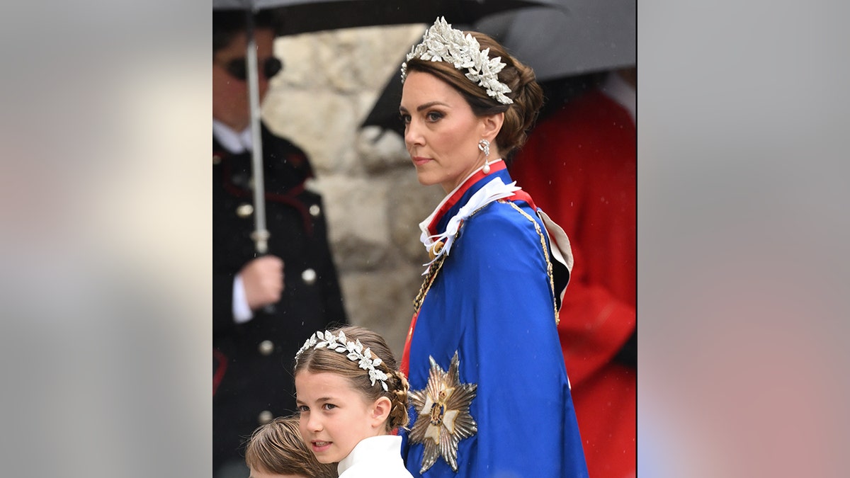 Kate Middleton in a leaf tiara walks in with a stoic expression to Westminster Abbey, wearing a blue cape, walking alongside her is Princess Charlotte in a matching, smaller tiara