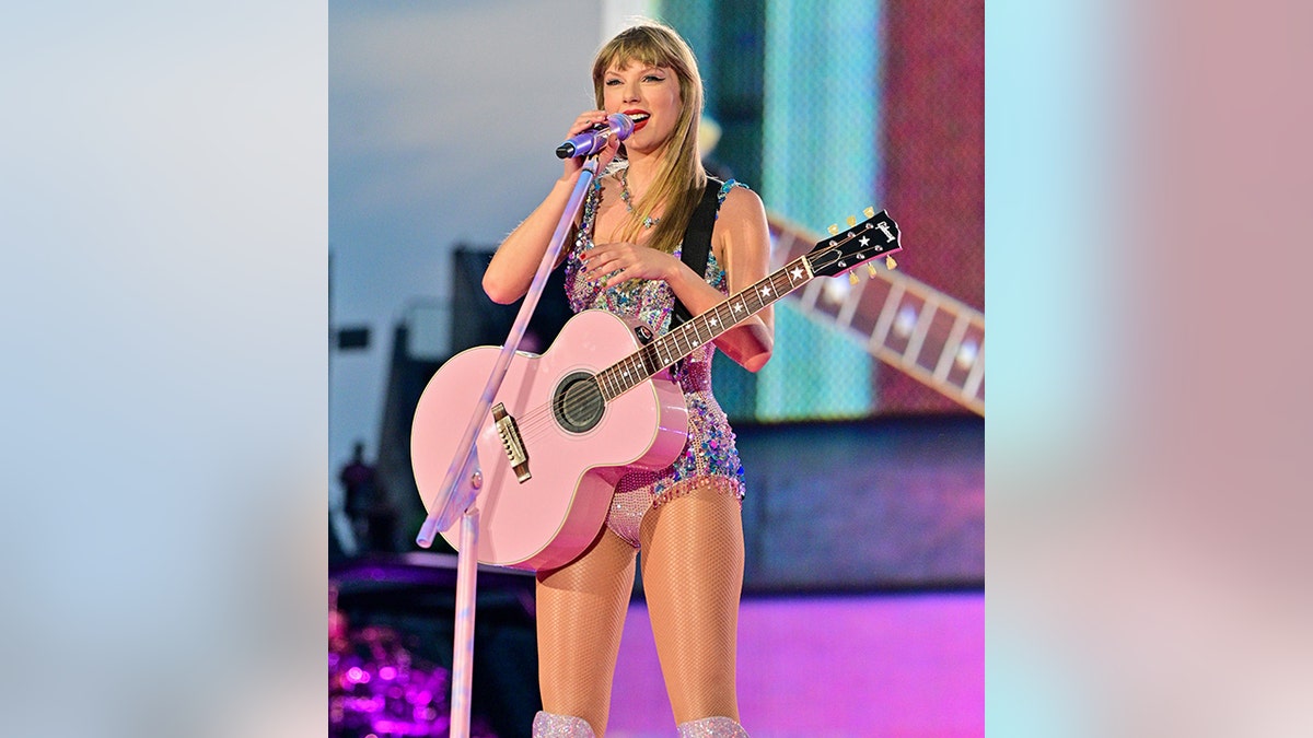 Taylor Swift holds the microphone during the Eras Tour playing on a pink guitar