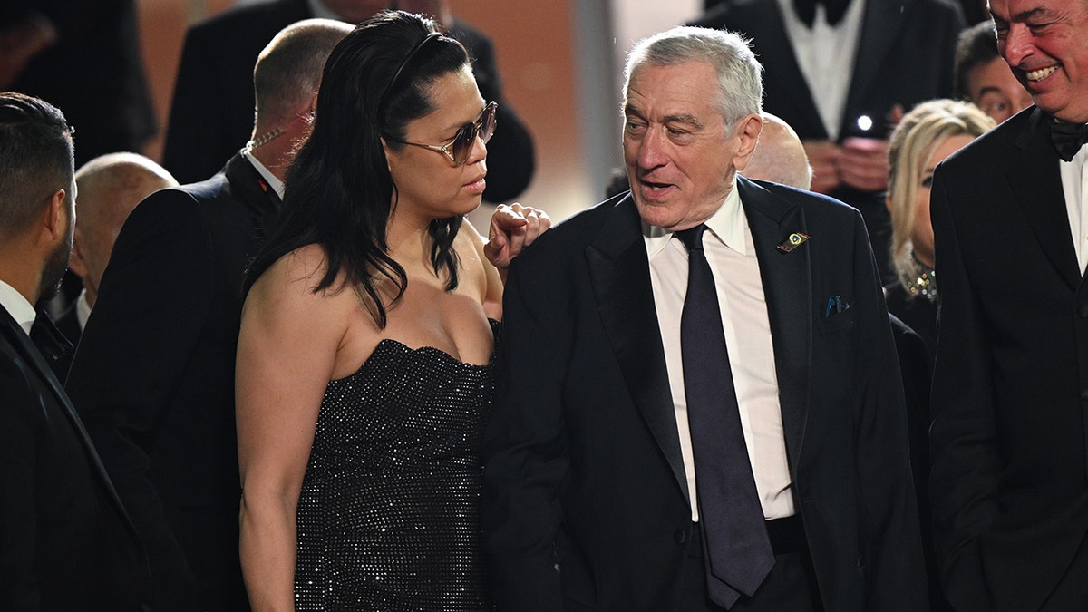 Tiffany Chen in a black sparkly gown and oversized sunglasses stands slightly behind Robert De Niro, who looks back to say something to her