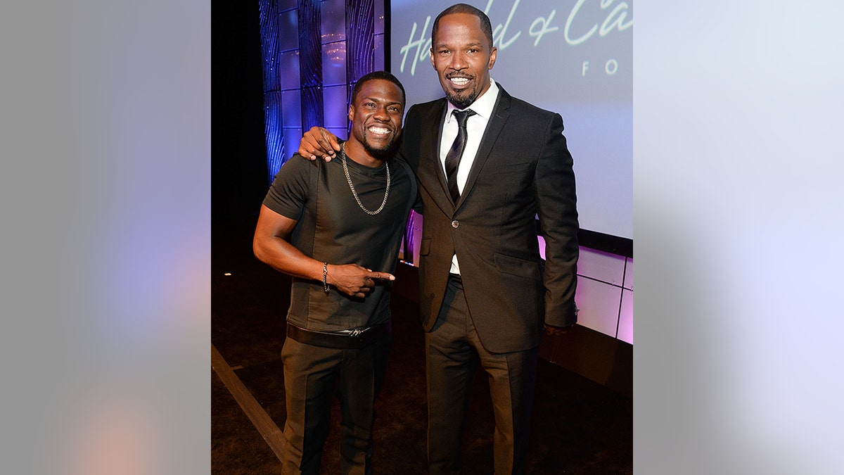 Kevin Hart wearing a black t-shirt and pants points to Jamie Foxx in a black suit and tie as they smile for a picture at an event