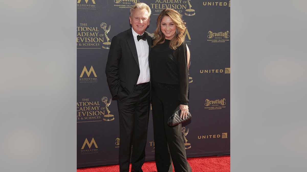 Pat Sajak in a black classic tuxedo smiles next to his wife with a black gown with slits on the sleeves