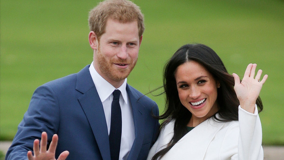 Prince Harry in a blue suit waves for photos as he walks out with his new fiancée Meghan Markle, wearing white and putting her hand up in the air