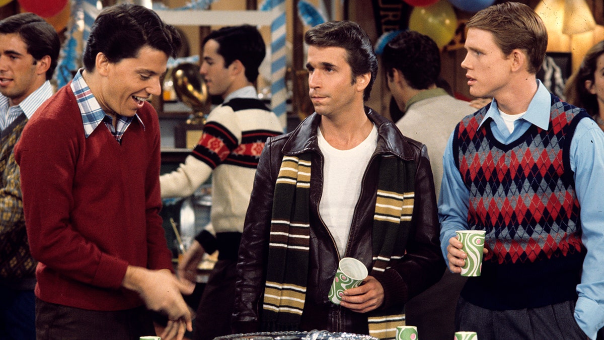 Anson Williams as Potsie, Henry Winkler as Fonzie, and Ron Howard as Richie hold cups at a party while getting something to drink in an episode of Happy Days, with Winkler wearing his character's signature leather jacket and a striped scarf