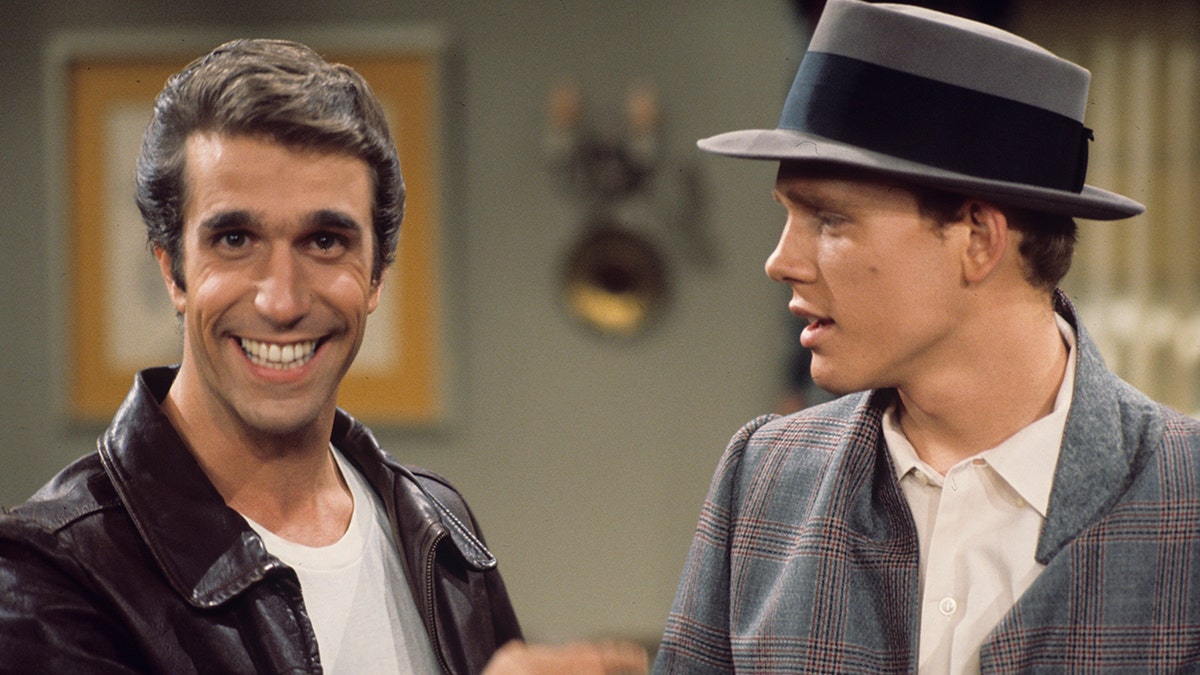 Henry Winkler as Fonzie stares at the camera and flashes a smile as Fonzie in a white shirt and leather jacket as Ron Howard as Richie stares at him wearing a brimmed hat in a season 3 episode of "Happy Days"