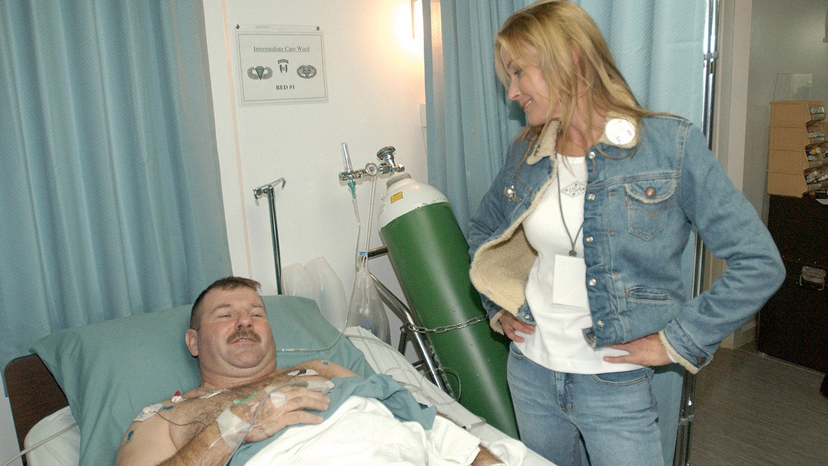 Bo Derek wearing a denim jacket and jeans with a matching white shirt speaking with a man lying on a hospital bed