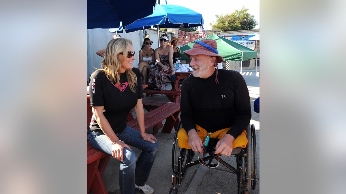 Bo Derek in a black outfit and sunglasses chatting with a man in a matching black outfit and a colorful hat