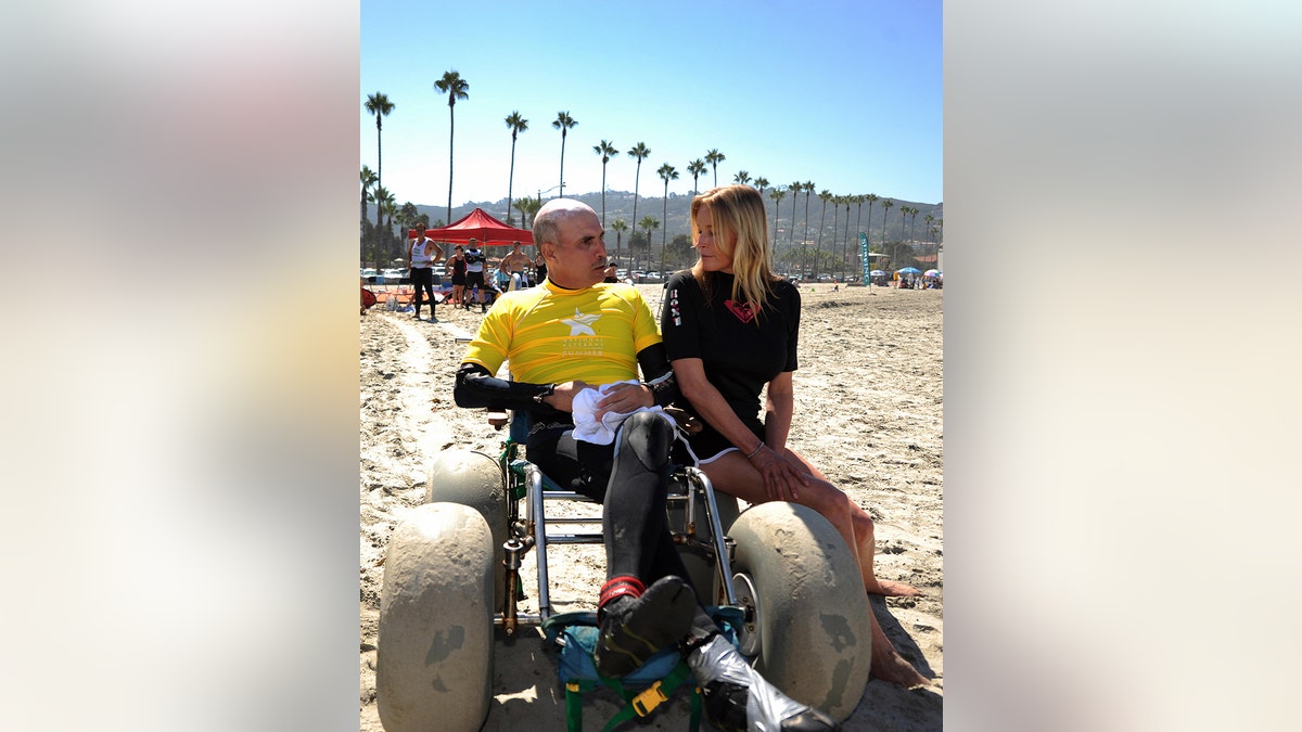 A disabled veteran wearing a yellow shirt in a wheelchair chatting with Bo Derek in a black outfit at the beach