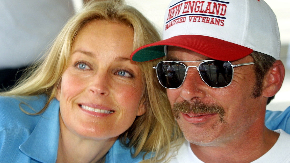 Bo Derek in a blue shirt leaning against a man with sunglasses and a red and white hat