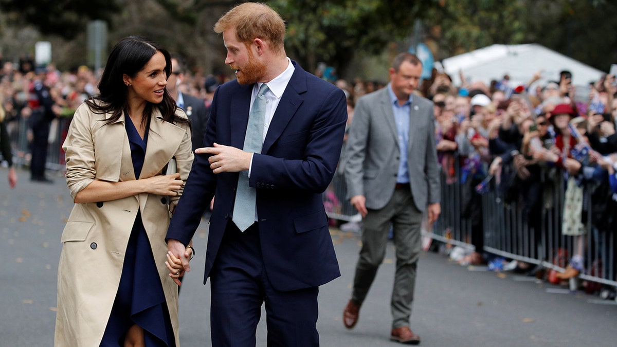 Meghan Markle wearing a trench coat and a black dress with prince harry wearing a dark suit and a light blue tie