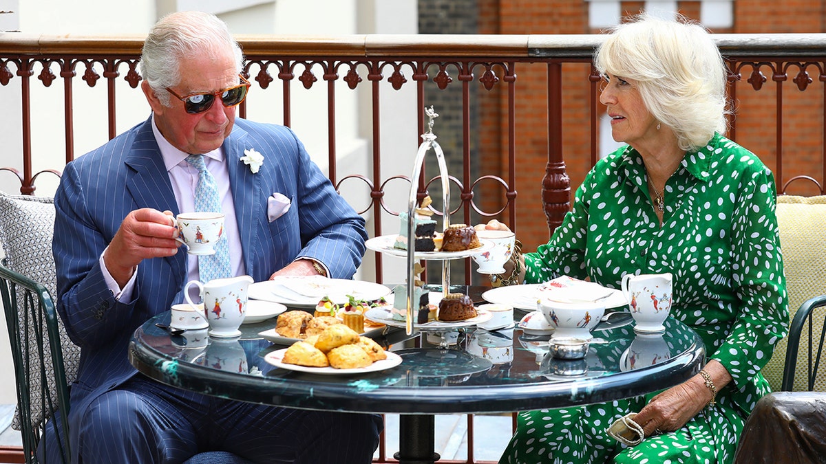King Charles in a light blue suit sitting with Queen Camila in a green and white dress as they have tea