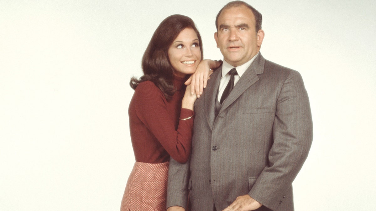 Mary Tyler Moore wearing a brown sweater and matching skirt leaning into Ed Asner in a grey suit