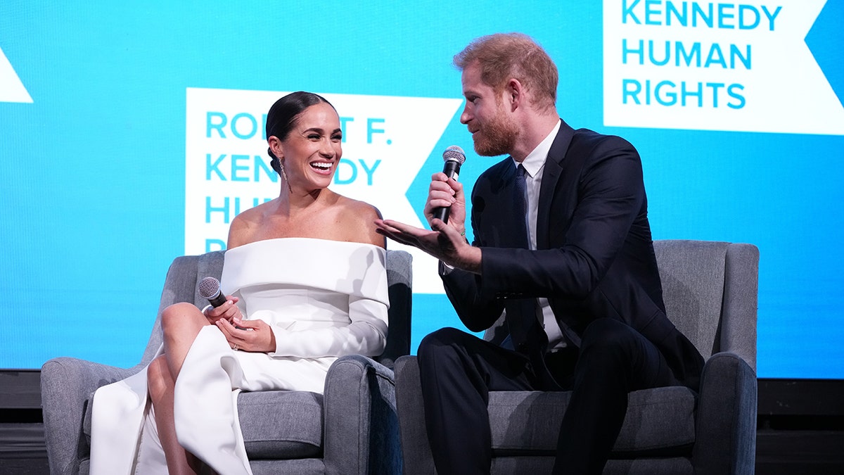 Meghan Markle wearing a white off the shoulder dress sitting next to Prince Harry in a suit and tie