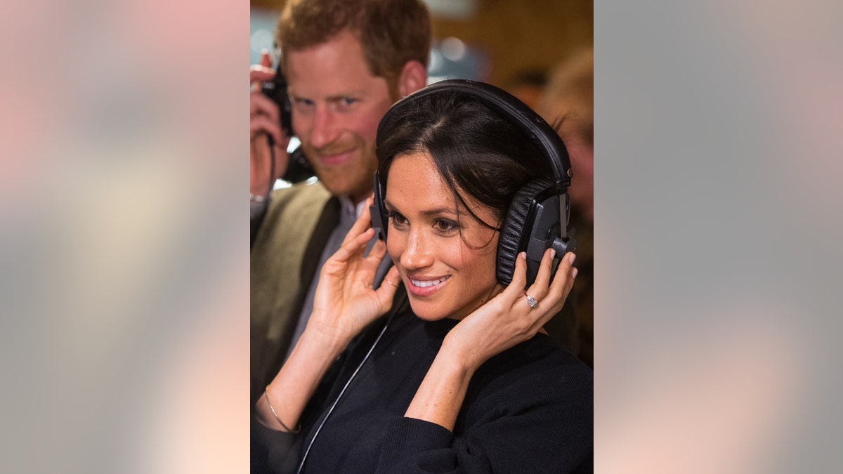 Meghan Markle in a black dress smiling and holding her headphones