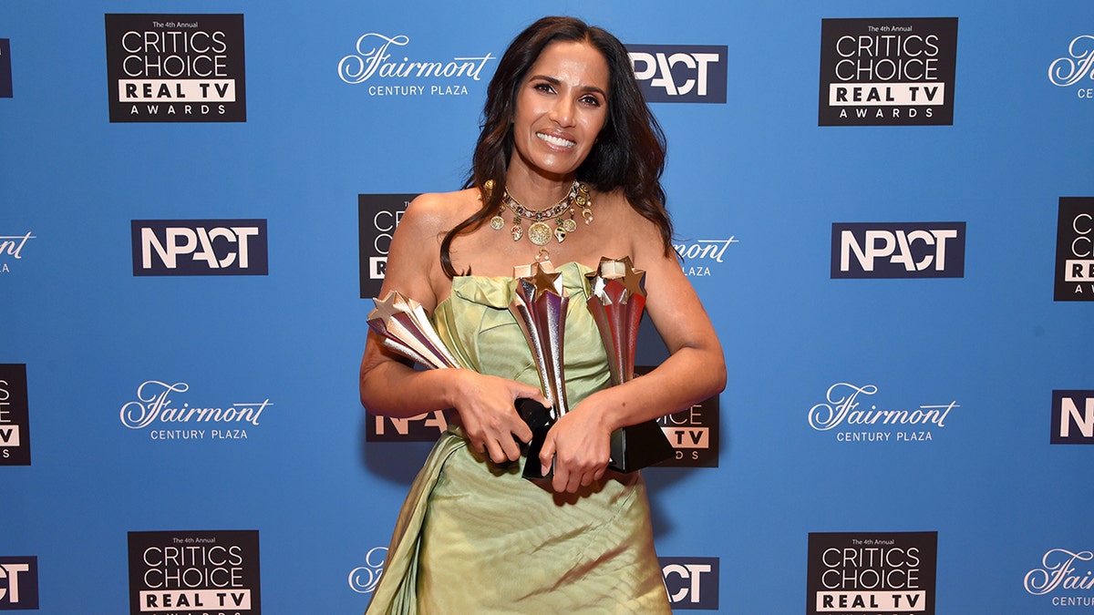 Padma Lakshmi wearing a light dress sleeveless dress holding a bunch of awards