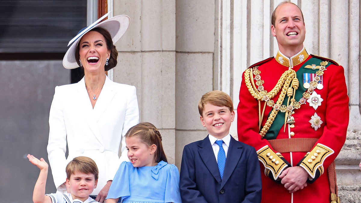 Kate Middleton wearing a white dress and a matching hat standing next to Prince William in a red royal uniform and their three children