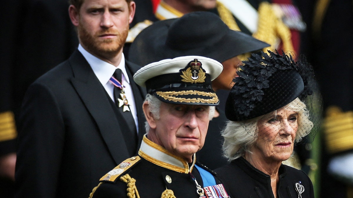 Prince Harry in a black suit, King Charles in his military uniform and Camilla in a black dress looking in the same direction