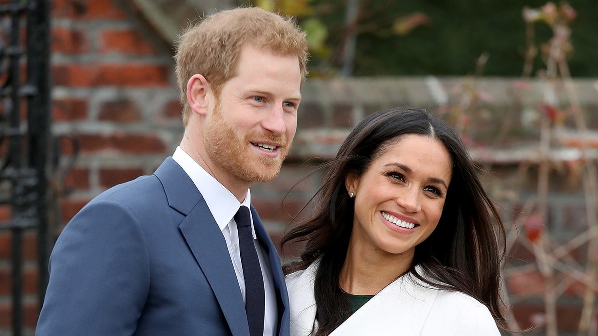 Meghan Markle wearing a white coat and a black dress next to Prince Harry in a blue suit and skinny black tie