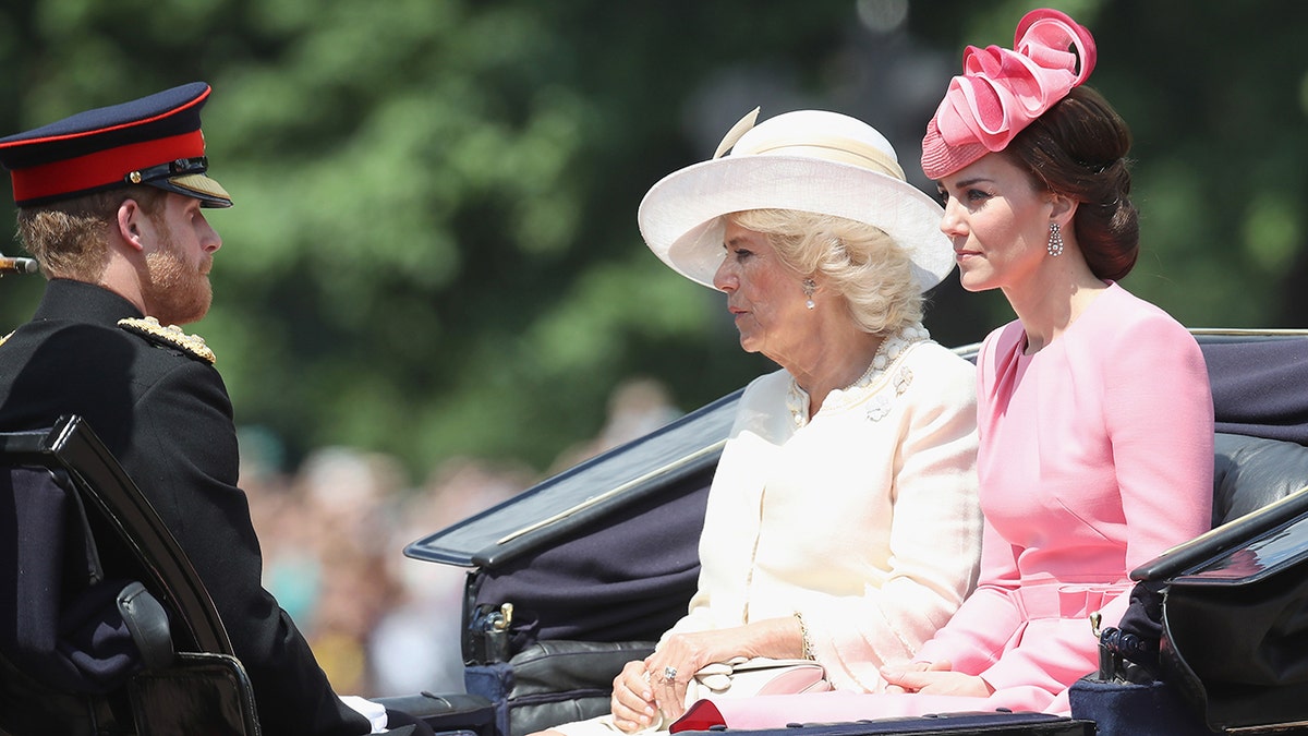 Prince Harry in his military uniform sitting across from Camilla in a white dress and matching hat and Kate Middleton in a pink dress and a matching hat