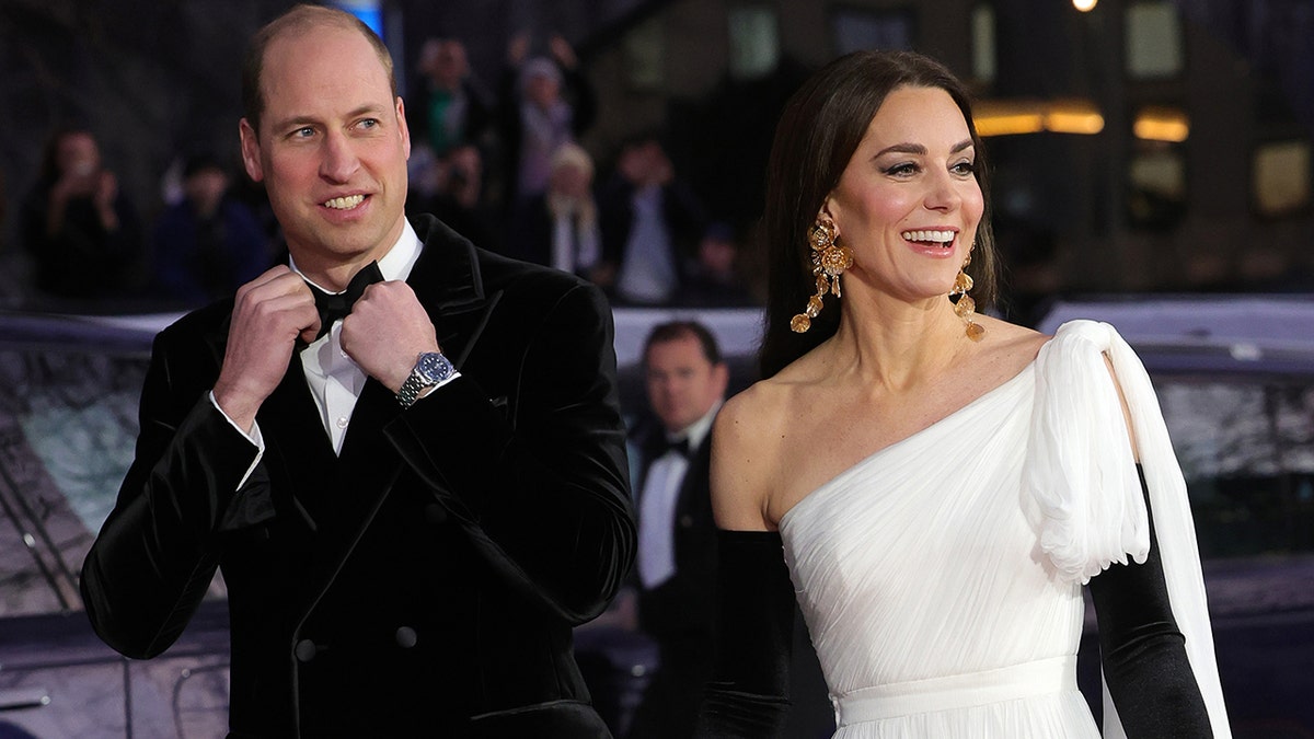 Prince William in a tux adjusting his bowtie walking next to Kate Middleton in a strapless white gown and black opera gloves
