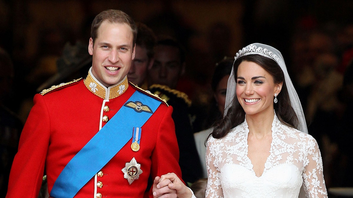 Prince William in a red royal uniform holding the hand of kate middleton wearing a bridal gown