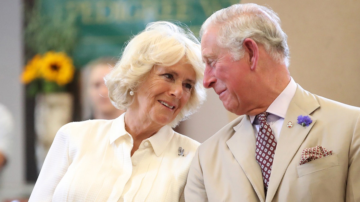 Queen Camilla in a beige blouse looks lovingly at her husband King Charles in a matching suit and tie