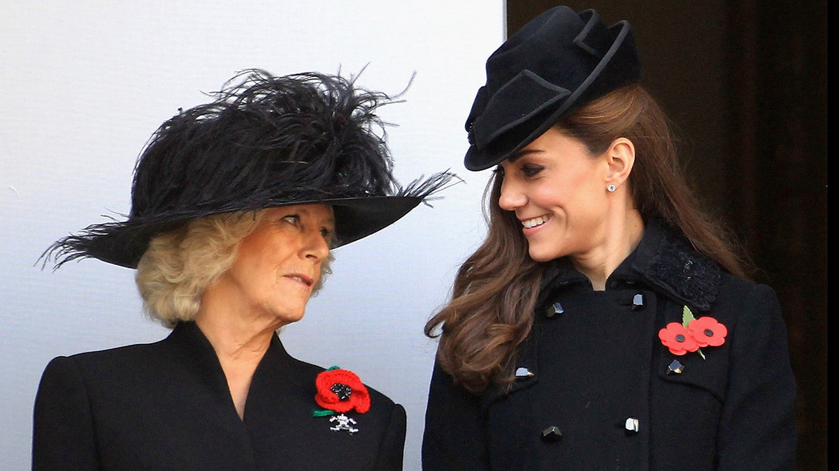 Queen Camilla in a black dress and matching hat looking at a smiling Kate Middleton also wearing a black coat and matching dress