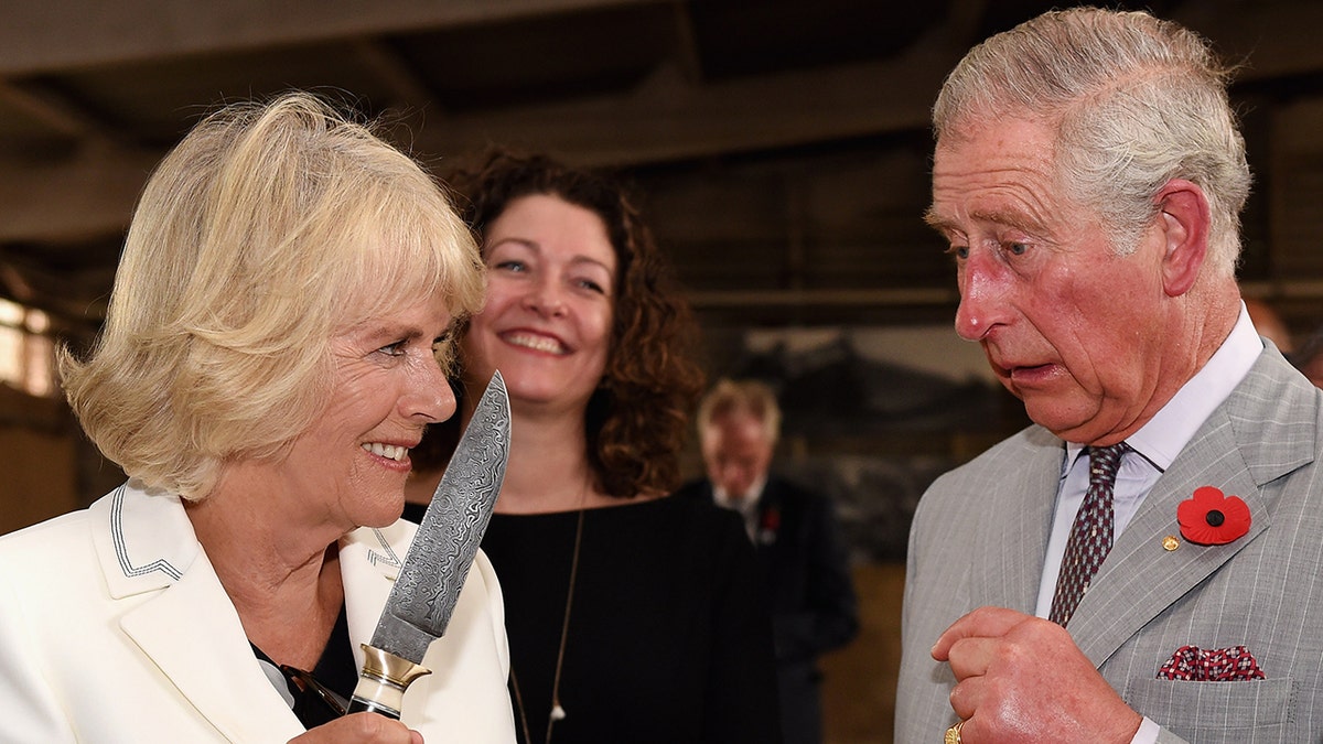 Camilla in a white blazer jokingly holding a knife as king charles in a grey suit looks on
