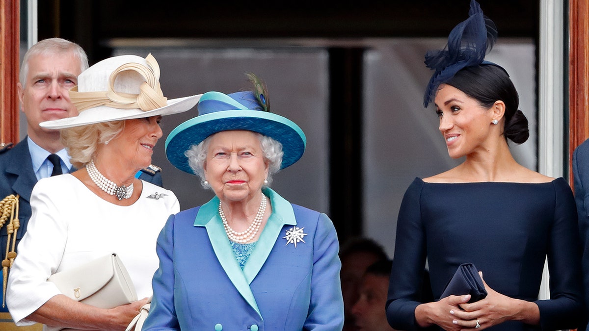 Camilla wearing a white dress chatting with Meghan Markle in a dark blue dress as Queen Elizabeth, wearing a multilayered blue dress, looks straighforward