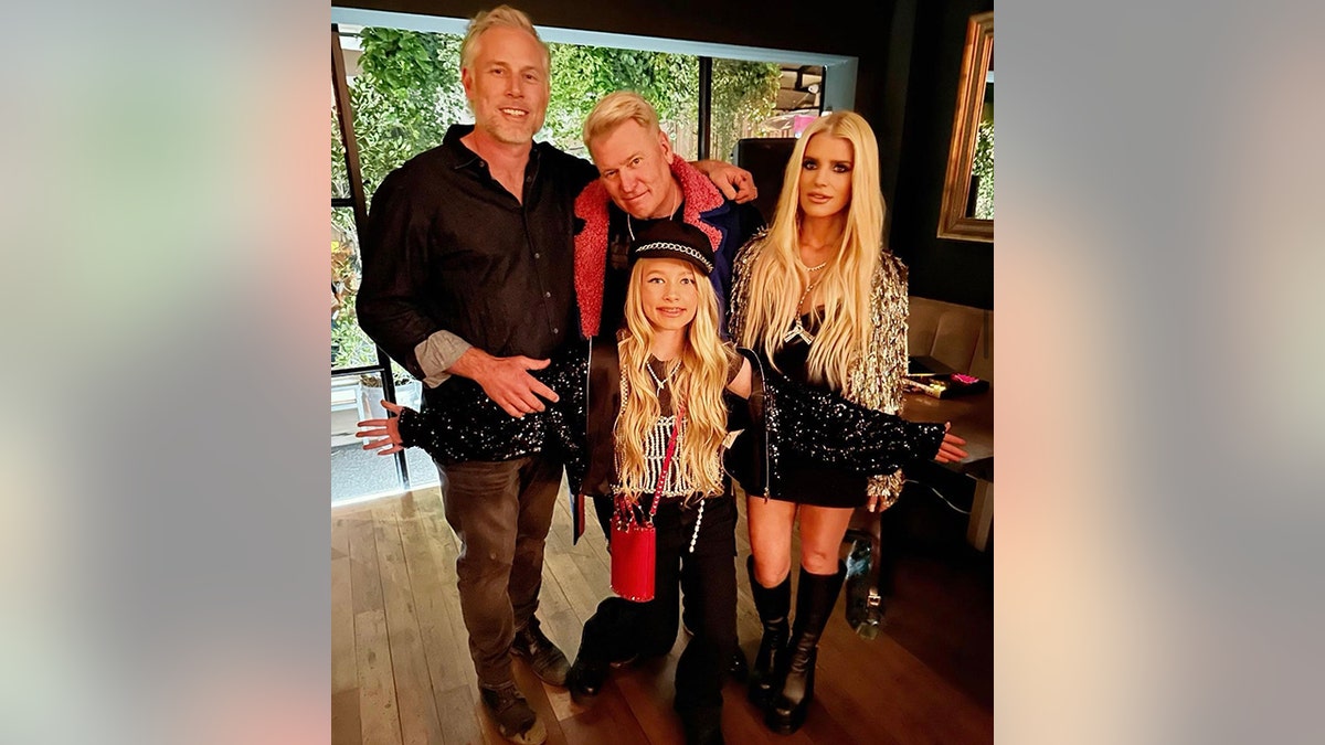 Eric Johnson in a black shirt and dark pants stands next to Joe Simpson, who is partially hunched wearing a pink furry jacket, next to Jessica Simpson in a mini skirt and sparkly gold top, and her daughter Maxwell Drew with a sparkly top in the front with her arms spread out