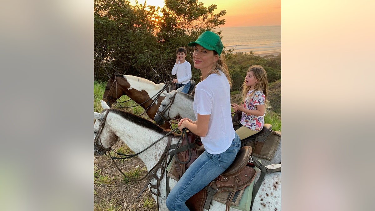 Gisele Bündchen looks back at a photo while riding on a horse with her children Benny and Vivi in the back, also on horses and a sunset in the background 