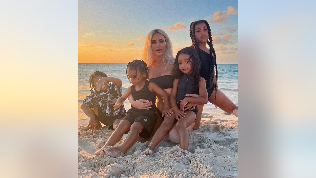 Kim Kardashian sits on the beach with her four children, balancing Chicago and Psalm on her laugh with North and Saint next to her