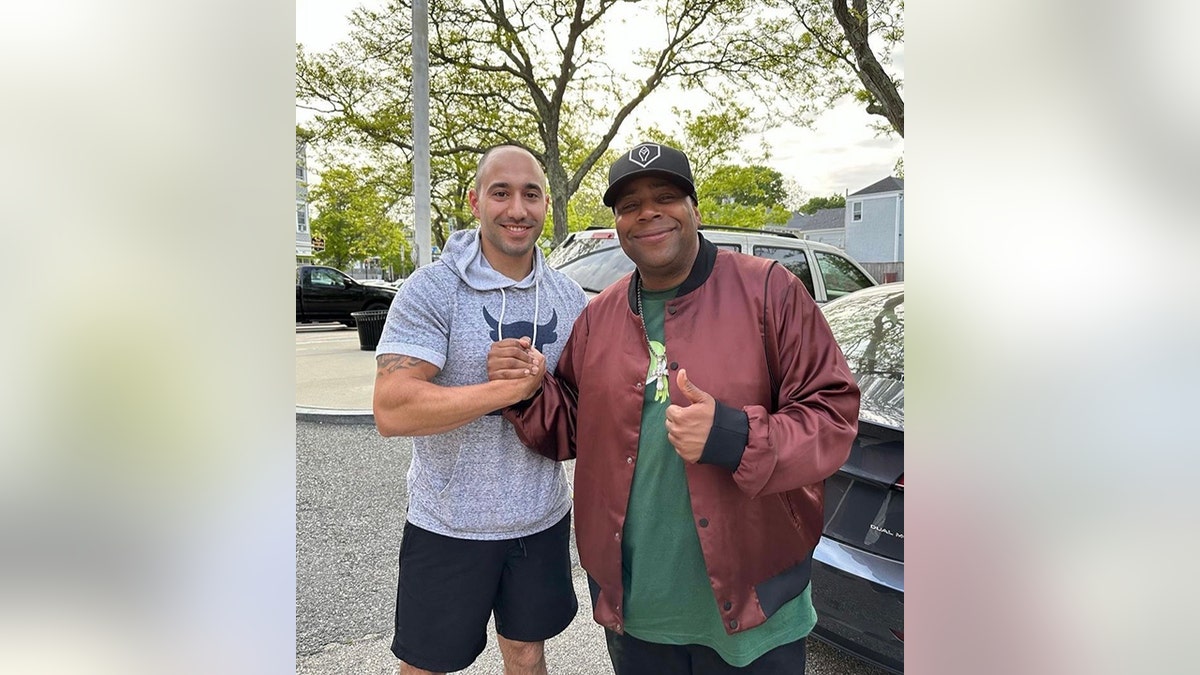 Kenan Thompson in a red jacket and green shirt holds hands with Community Officer Frank Pirri