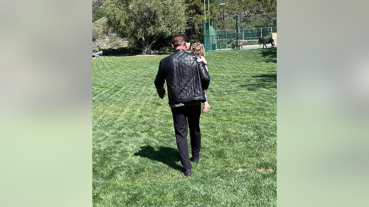 Arnold Schwarzenegger walks in black with his back to the camera holding granddaughter Lyla