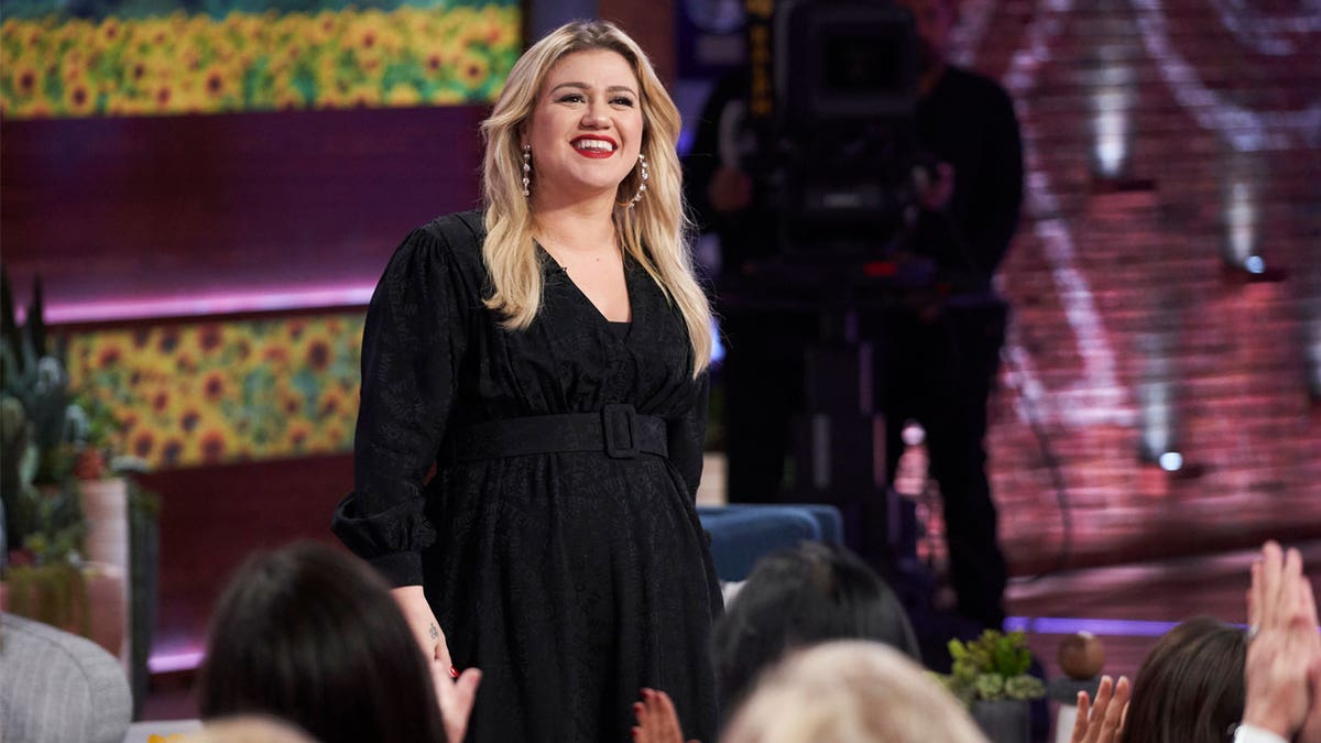 Kelly Clarkson smiles in a black dress on her talk show while looking out to the audience