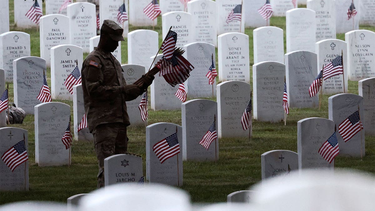 Military member holding flags and placing them on a tombstone.