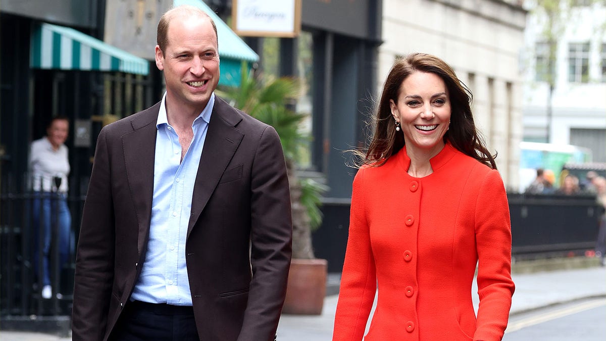 Príncipe William com camisa azul e casaco marrom caminha ao lado de Kate Middleton com casaco longo vermelho/laranja brilhante