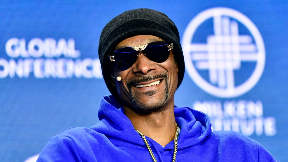 Snoop Dogg in a blue sweatshirt and black hat smiles on stage with black sunglasses on