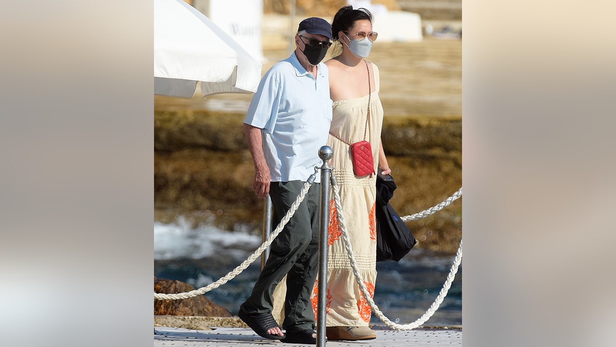 Robert De Niro wears a black cap and facemask and walks on a boardwalk in France with Tiffany Chen, in a long dress, also wearing a facemask