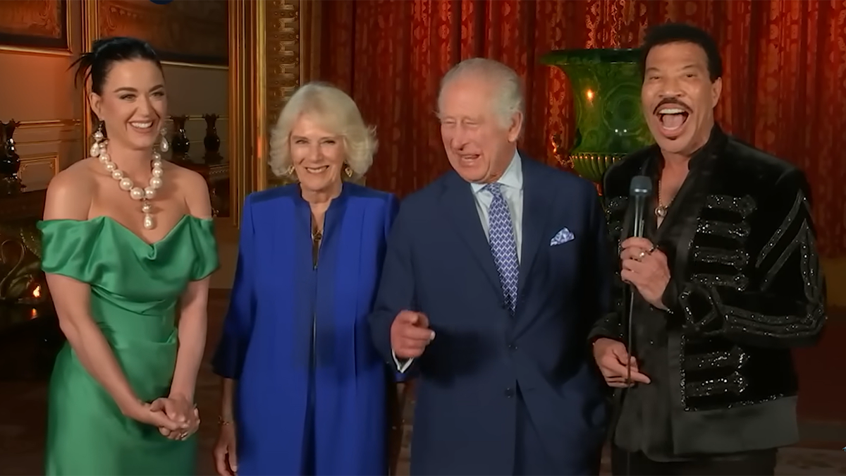 Katy Perry chuckles in a green gown with sleeves on the side and a massive pearl necklace next to Queen Camilla in a royal blue suit, next to King Charles in a navy suit and blue patterned tie next to Lionel Richie in black, holding a microphone