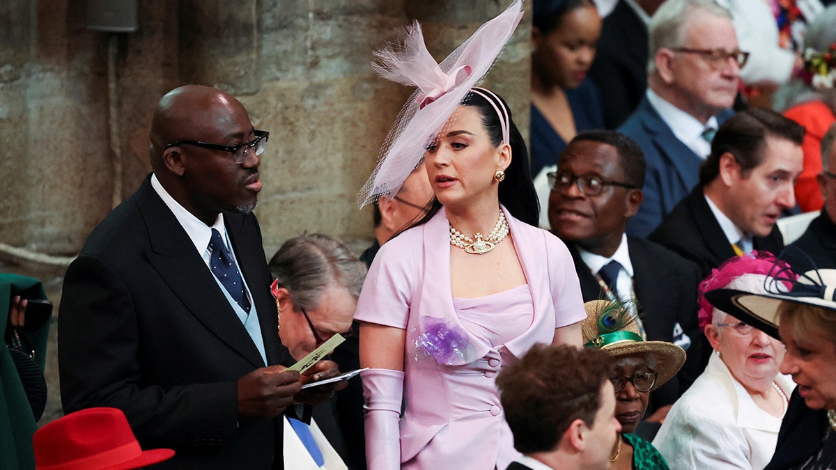 Katy Perry in a lilac dress and large headband hat struggles to find her seat in Westminster Abbey