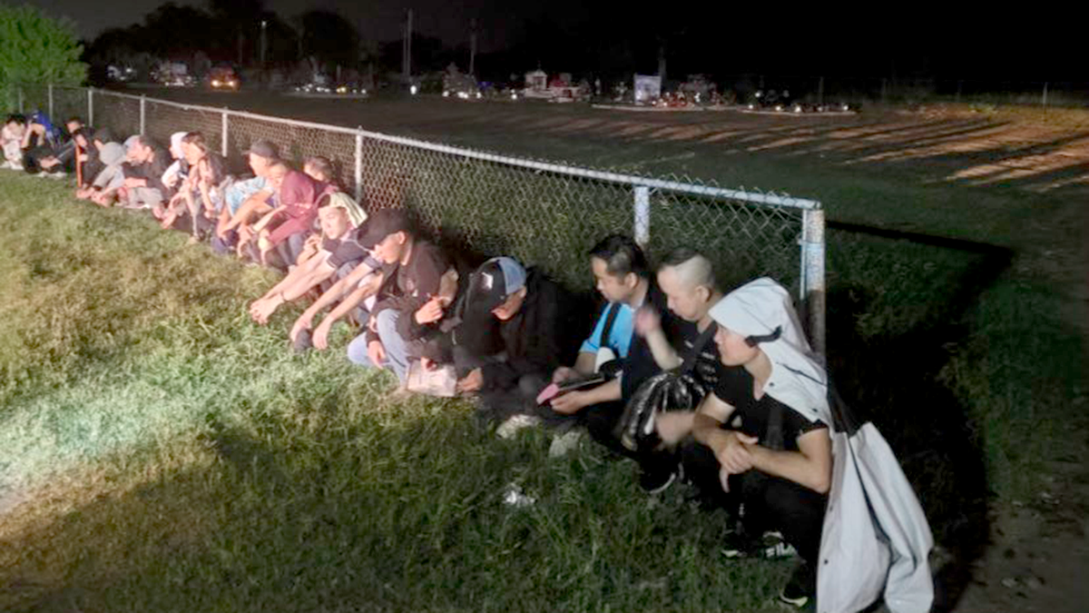 A line of migrants sat along a fence at night