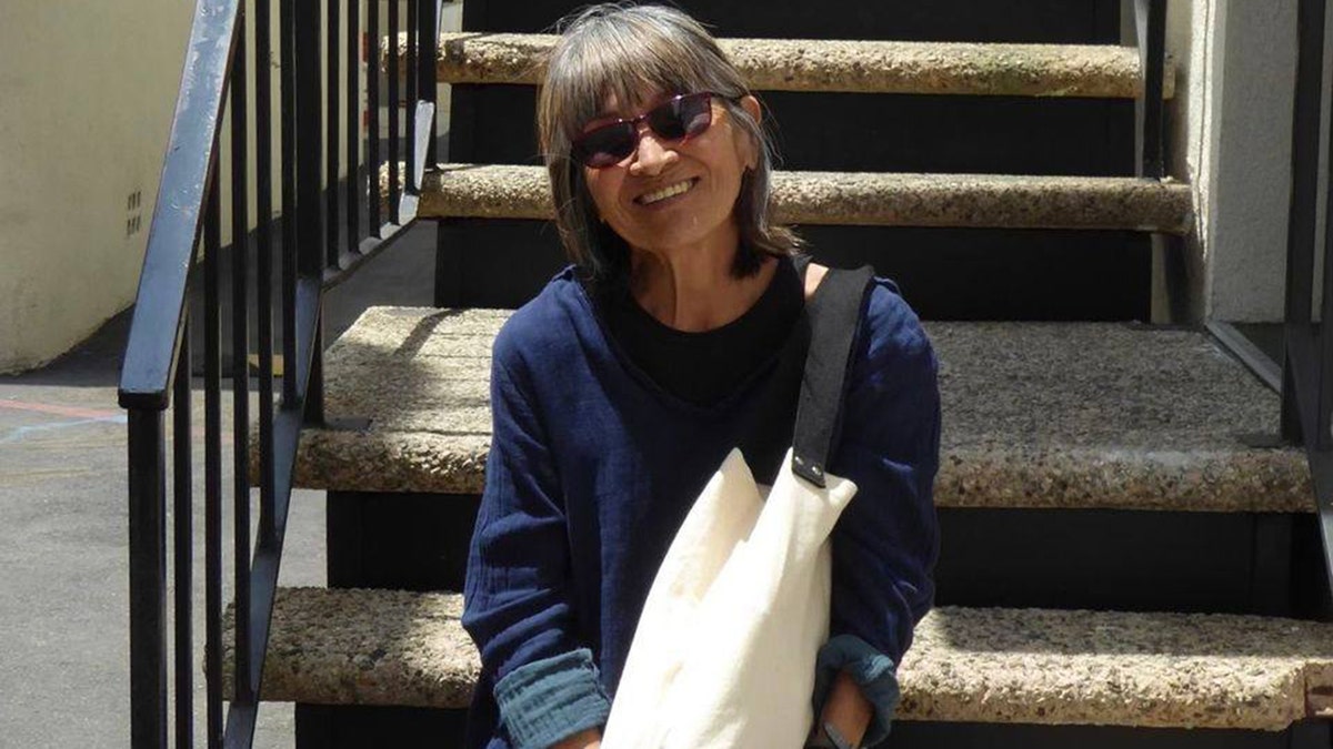 MASH star Eileen Saki wears blue sweater and carries canvas tote while perched on staircase