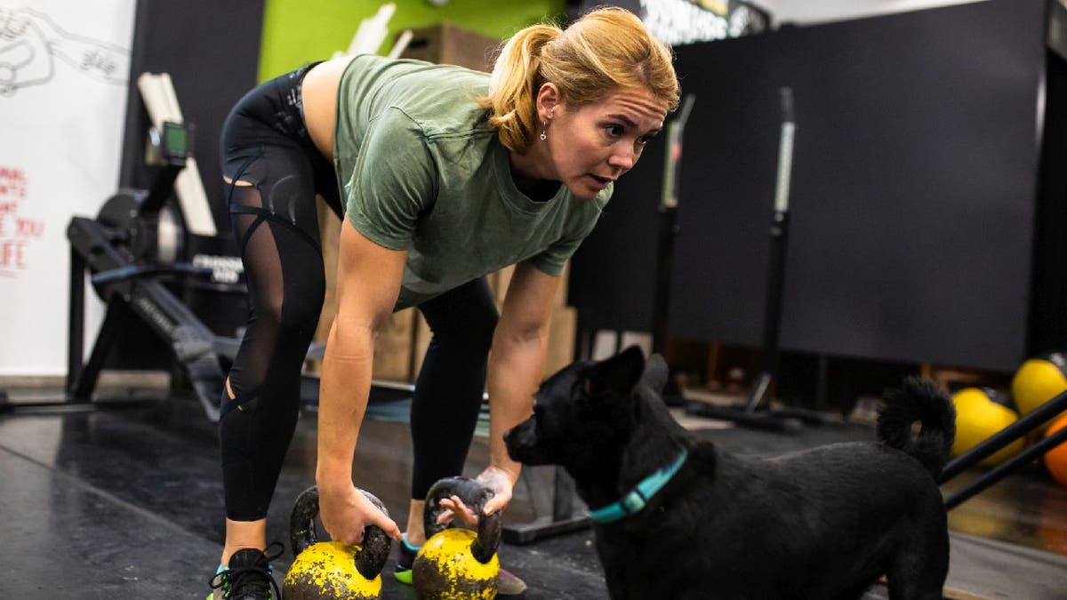 A woman deadlifts with kettlebells in a gym while her dog watches.