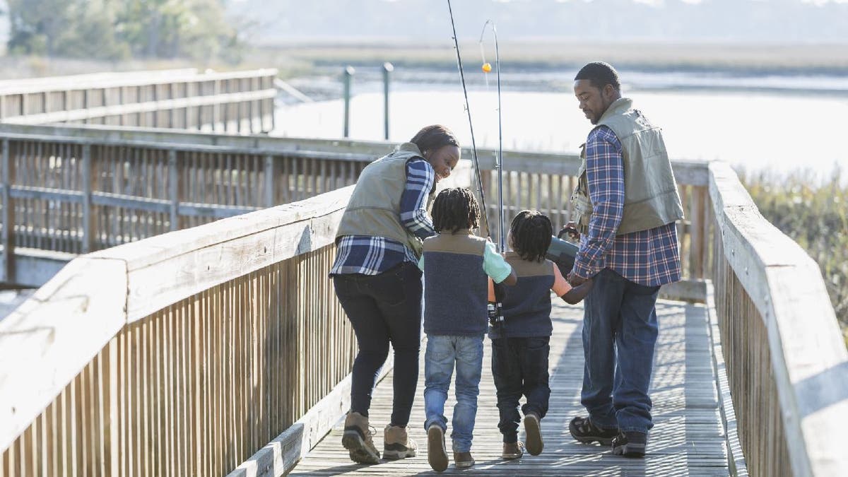 Rear view of a young family with two boys going fishing. They are walking on a wooden walkway or bridge, carrying rods and a tackle box.